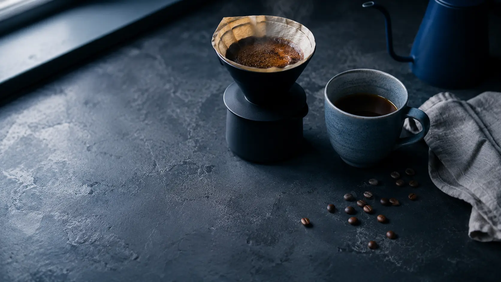 Pour-over coffee dripper and mug on a dark charcoal-blue countertop, steam rising