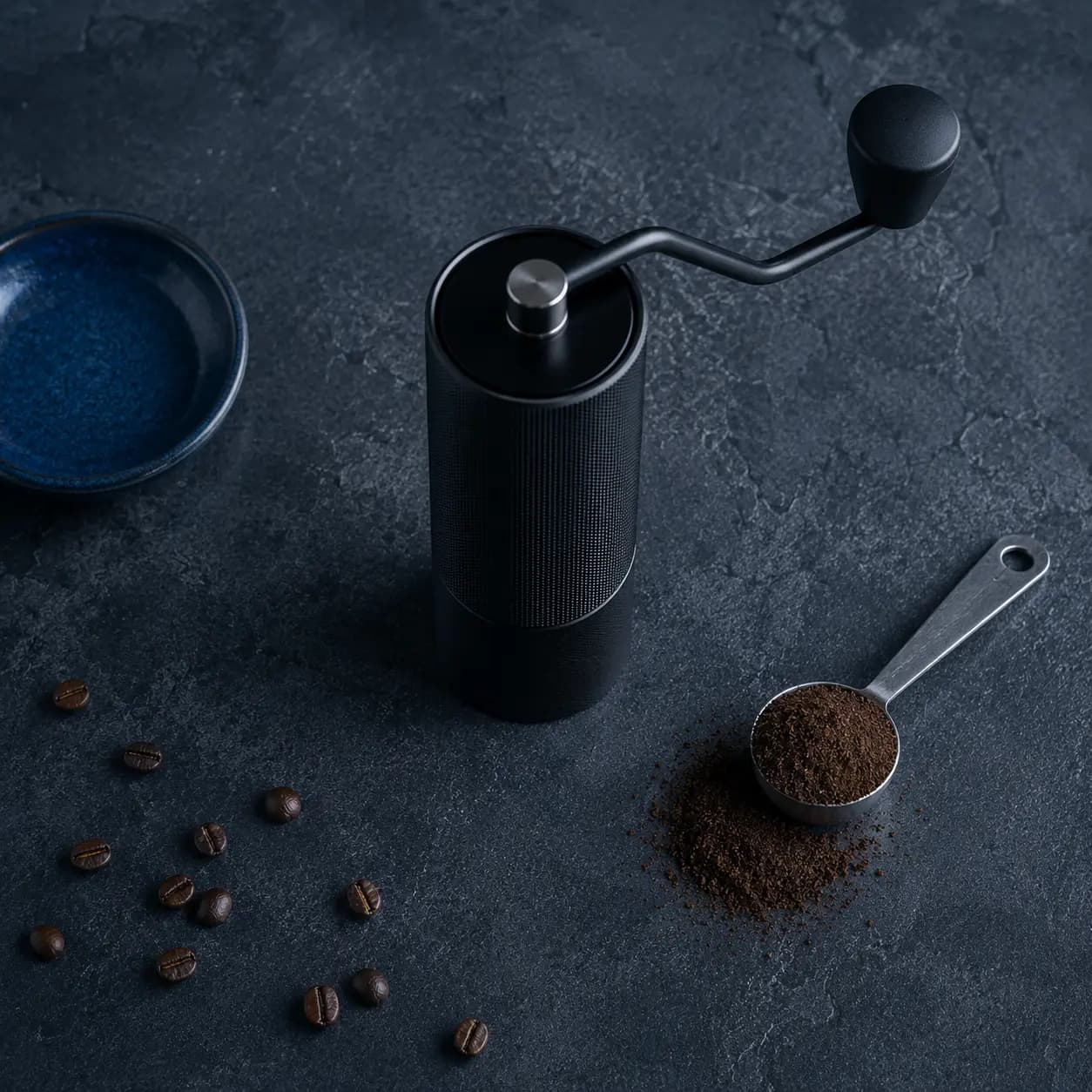 Hand coffee grinder and a scoop of fresh grounds on a dark charcoal-blue surface, top-down
